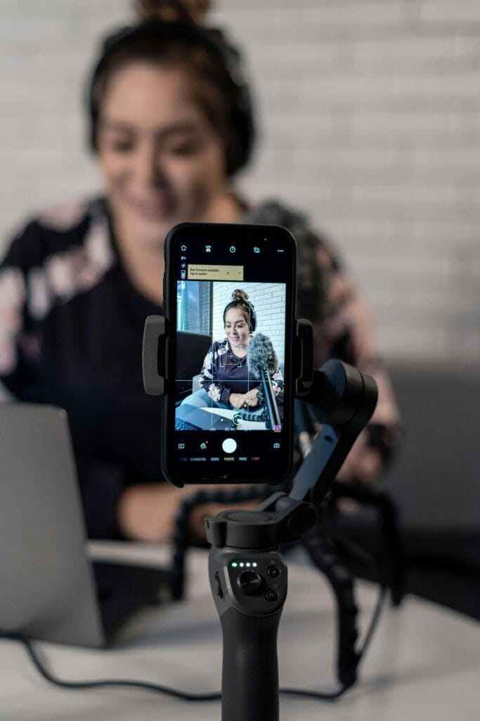 Smartphone on a tripod recording a smiling woman wearing headphones, microphone in foreground.