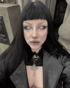 Gothic portrait of a person with black hair and bangs, pale makeup, facial piercings, wearing a black leather jacket and an ornate cross necklace outdoors near stone architecture.