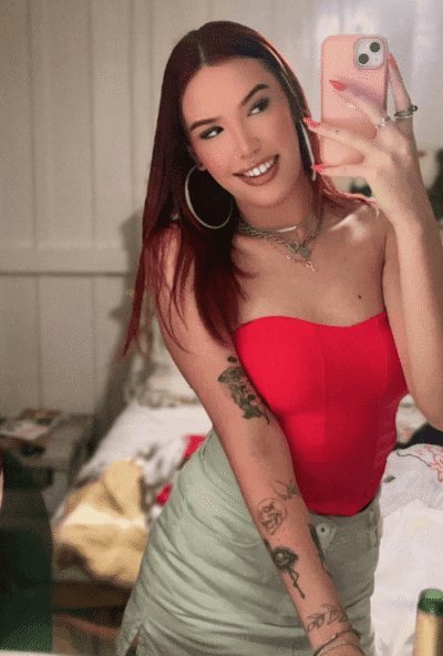 Woman taking a mirror selfie, wearing a red strapless top, light green skirt, hoop earrings, and layered necklaces, with visible arm tattoos.