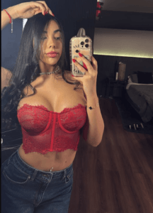 Woman in red lace bustier takes a mirror selfie in a bedroom, holding her phone up to camera.
