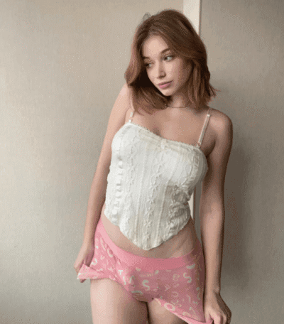 Woman standing against a light wall, wearing a white lace top and pink shorts, looking to her left with a relaxed pose.