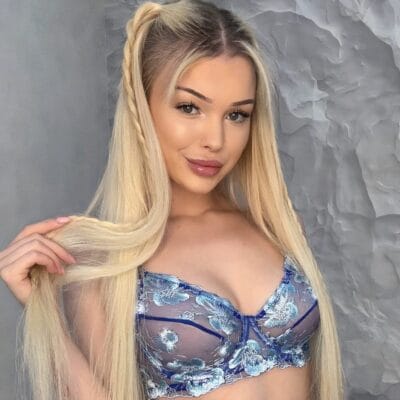 Young woman with long blonde hair in a blue floral lace bralette, facing the camera against a grey textured wall.