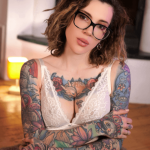 Portrait of a tattooed woman with short wavy hair and black-framed glasses, wearing a white lace top, facing the camera in a warmly lit indoor setting.